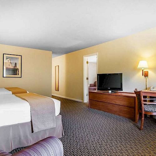 A hotel room with a bed, a TV on a wooden dresser, a desk with chairs, a lamp, and framed artwork on the wall. The carpet is gray and patterned.