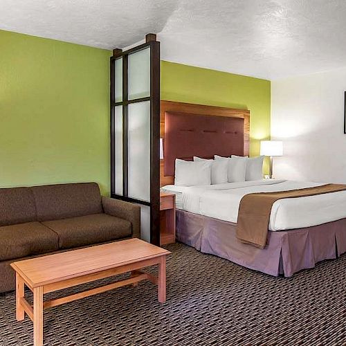 A hotel room with a bed, sofa, coffee table, bedside lamps, green accent wall, wall painting, and carpeting is shown.