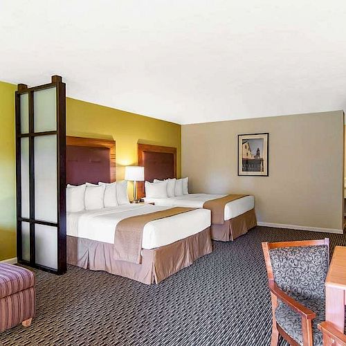 A hotel room with two beds, a TV, a desk, an armchair, and framed artwork on a green and beige wall, divided by a modern screen.