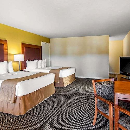 This image shows a hotel room with two double beds, a TV on a wooden dresser, a desk with a chair, and a lamp on the wall.