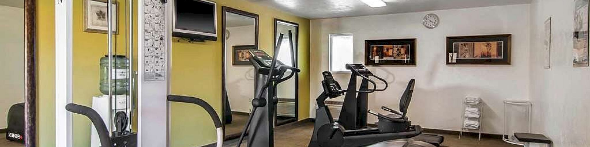 A small gym with various exercise machines, including ellipticals and a weight machine, bright lighting, and framed pictures on the walls.