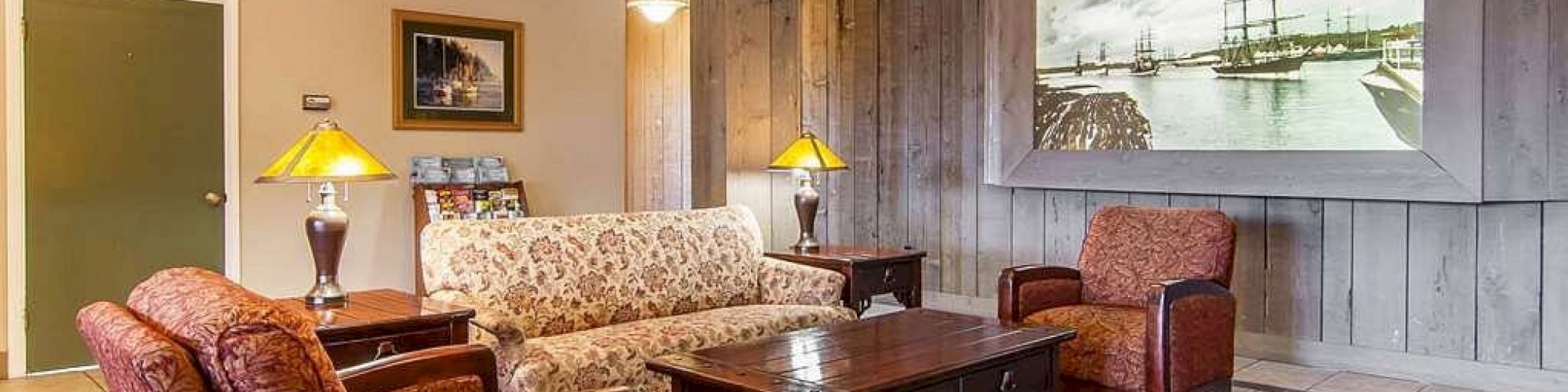 A cozy living room with a patterned sofa, two armchairs, wooden coffee table, side lamps, large wall art, and wooden paneling on one wall.