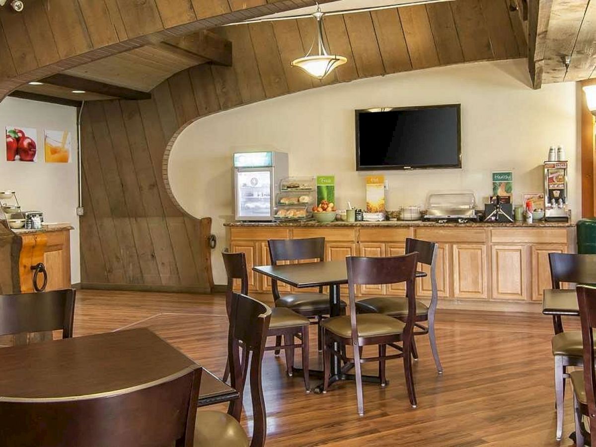 This image shows a cozy dining area with wooden decor, round tables, chairs, a TV, and a breakfast bar featuring various food items and beverages.