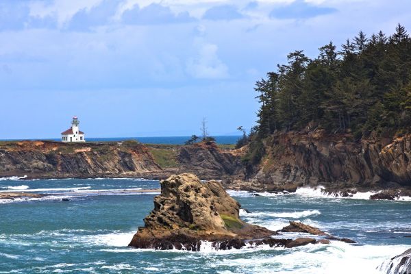 A lighthouse stands on a cliff overlooking the ocean with rocky shorelines and distant trees under a partly cloudy sky.