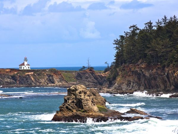 A lighthouse stands on a cliff overlooking the ocean with rocky shorelines and distant trees under a partly cloudy sky.