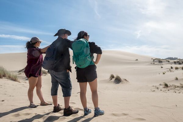 Three people stand in a sandy desert landscape looking into the distance, each wearing backpacks and hats under a blue sky.