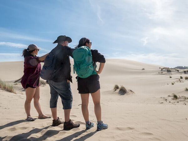 Three people stand in a sandy desert landscape looking into the distance, each wearing backpacks and hats under a blue sky.
