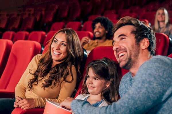 A group of people, including a family with a child, are sitting in a theater with red seats, smiling and looking at the screen, holding popcorn.