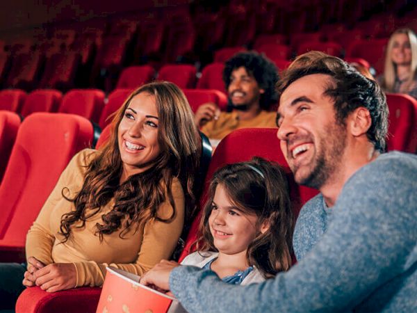 A group of people, including a family with a child, are sitting in a theater with red seats, smiling and looking at the screen, holding popcorn.
