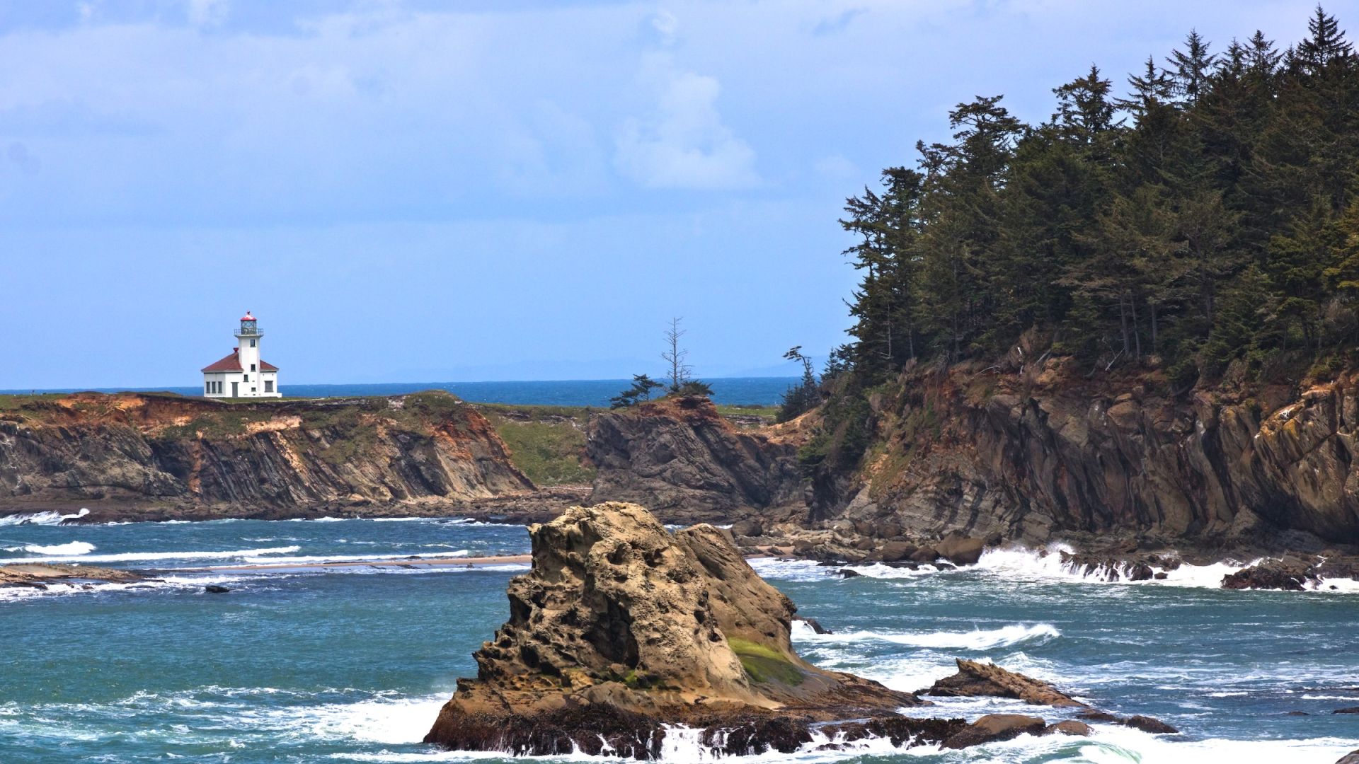 A scenic coastal view with a lighthouse atop a cliff, lush trees, rocky formations, and waves crashing against the shore ends the sentence.