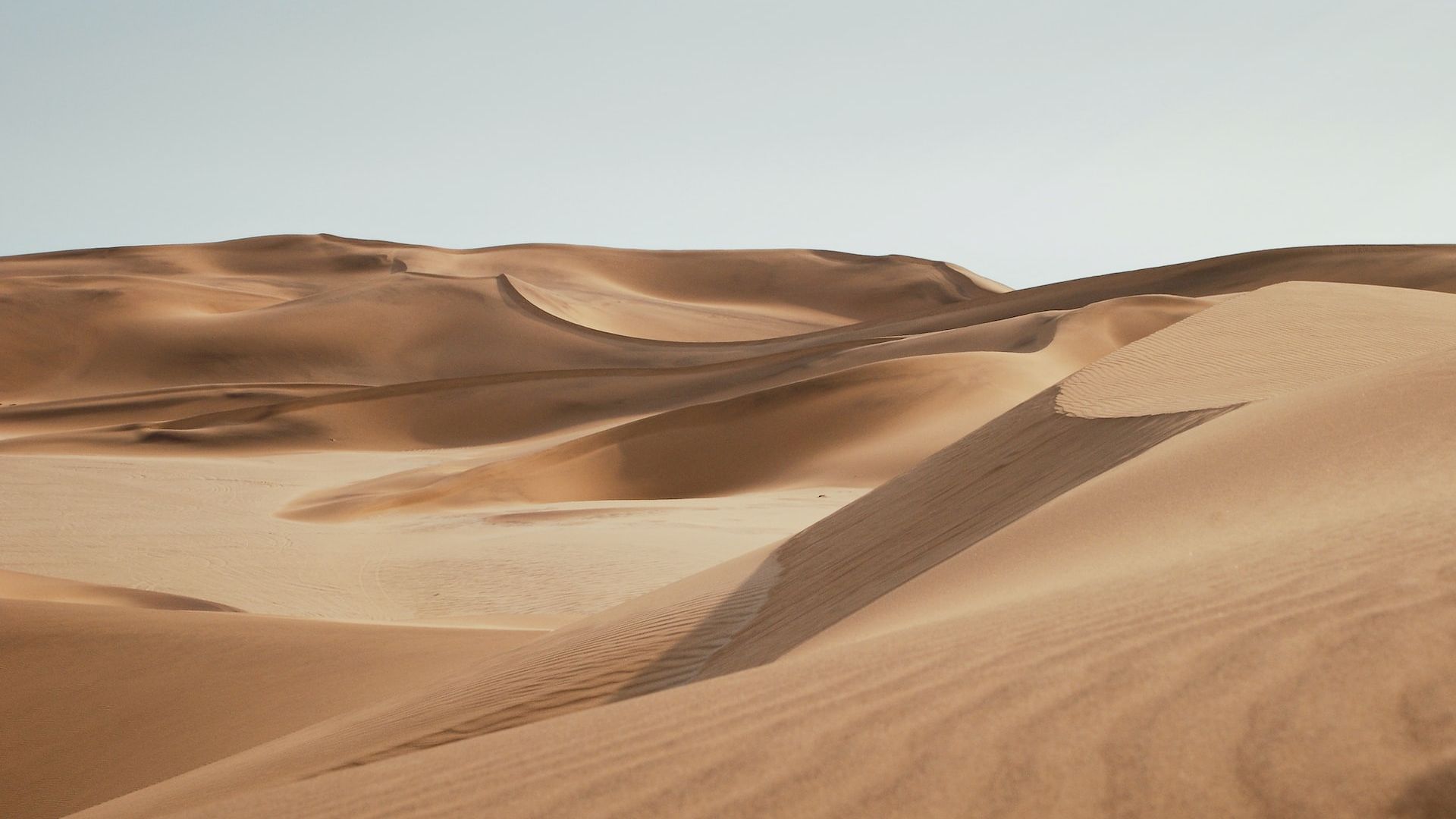 Sand dunes in a desert landscape with blue sky, showcasing shadow patterns and undulating shapes of sand formations, under natural sunlight.