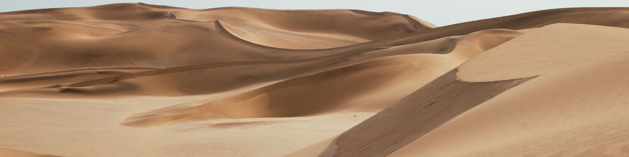 Sand dunes in a desert landscape with blue sky, showcasing shadow patterns and undulating shapes of sand formations, under natural sunlight.