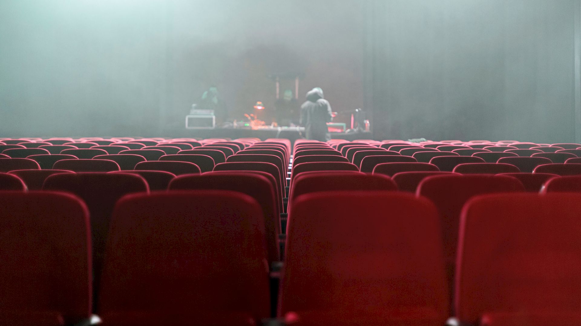 Rows of red theater seats with a hazy atmosphere, two people can be seen preparing the stage in the background.