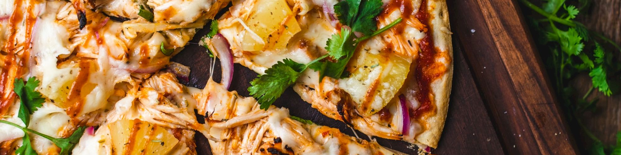 A sliced pizza topped with pineapple, red onions, shredded chicken, cilantro, and melted cheese on a wooden board, with cilantro scattered around.