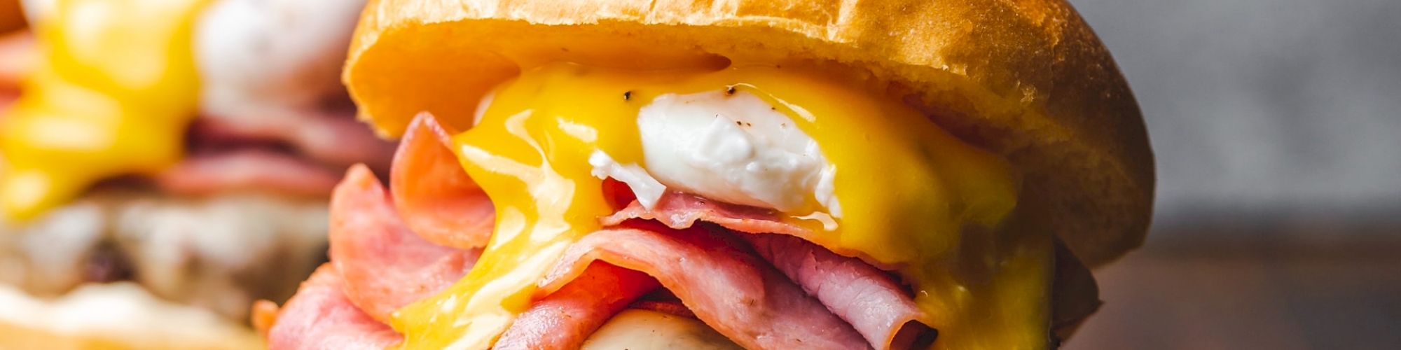 A juicy burger topped with cheese, ham, and a fried egg, with a knife inserted in the bun, sits on a wooden cutting board.