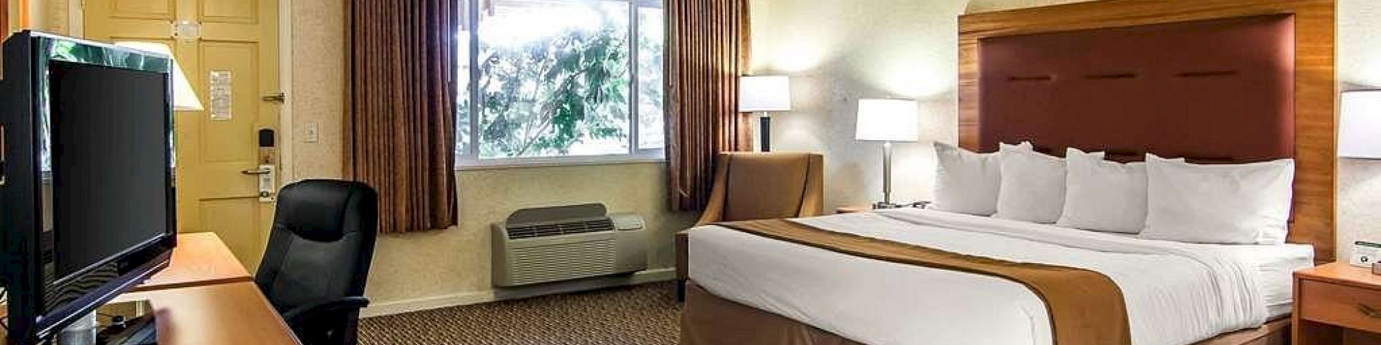 The image shows a hotel room with a large bed, a TV on a wooden desk, a chair, lamps, a window with curtains, and carpeted flooring.