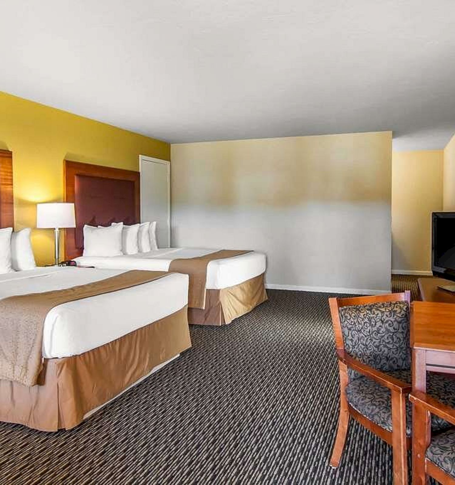 The image shows a hotel room with two double beds, a TV, a desk with a chair, a lamp, and a mirror, decorated in neutral tones.