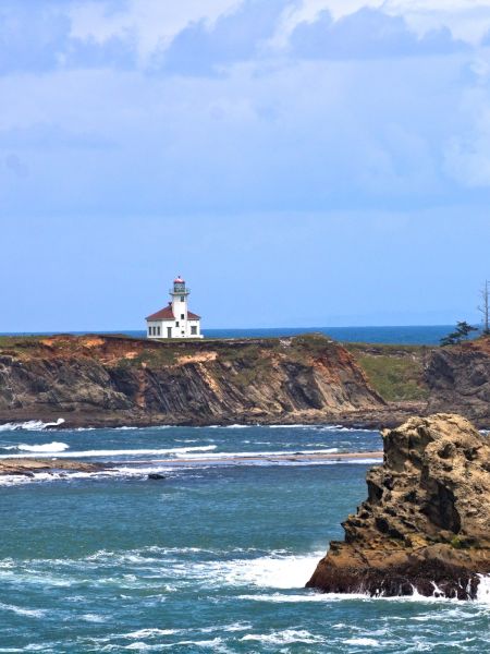 A scenic coastal view with a lighthouse on a distant cliff, rocky formations in the water, and dense trees on the right side.