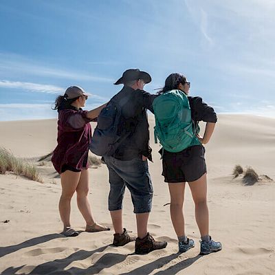 Three people standing in a desert environment, dressed in outdoor attire with backpacks, looking into the distance on a bright, sunny day.