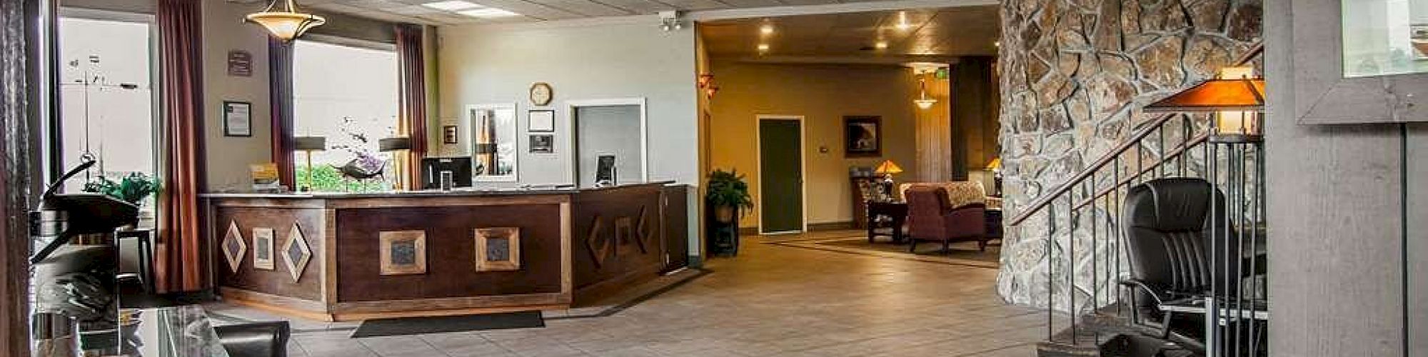 A hotel lobby with a reception desk, seating area, stone wall, and hanging lights. There are armchairs and a desk with a chair on the side.