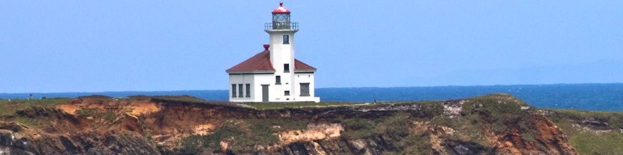 A lighthouse sits atop a rugged cliff overlooking the ocean, with waves crashing along the shoreline under a partly cloudy sky.