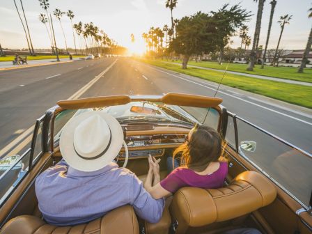 A couple is driving in a convertible on a palm-lined road at sunset, holding hands and enjoying the scenic view.