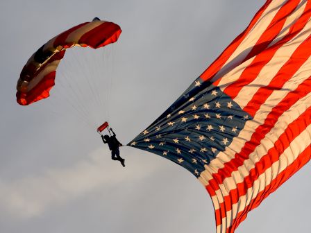 A parachutist is descending with an open parachute and holding a large American flag that is billowing in the air behind him.