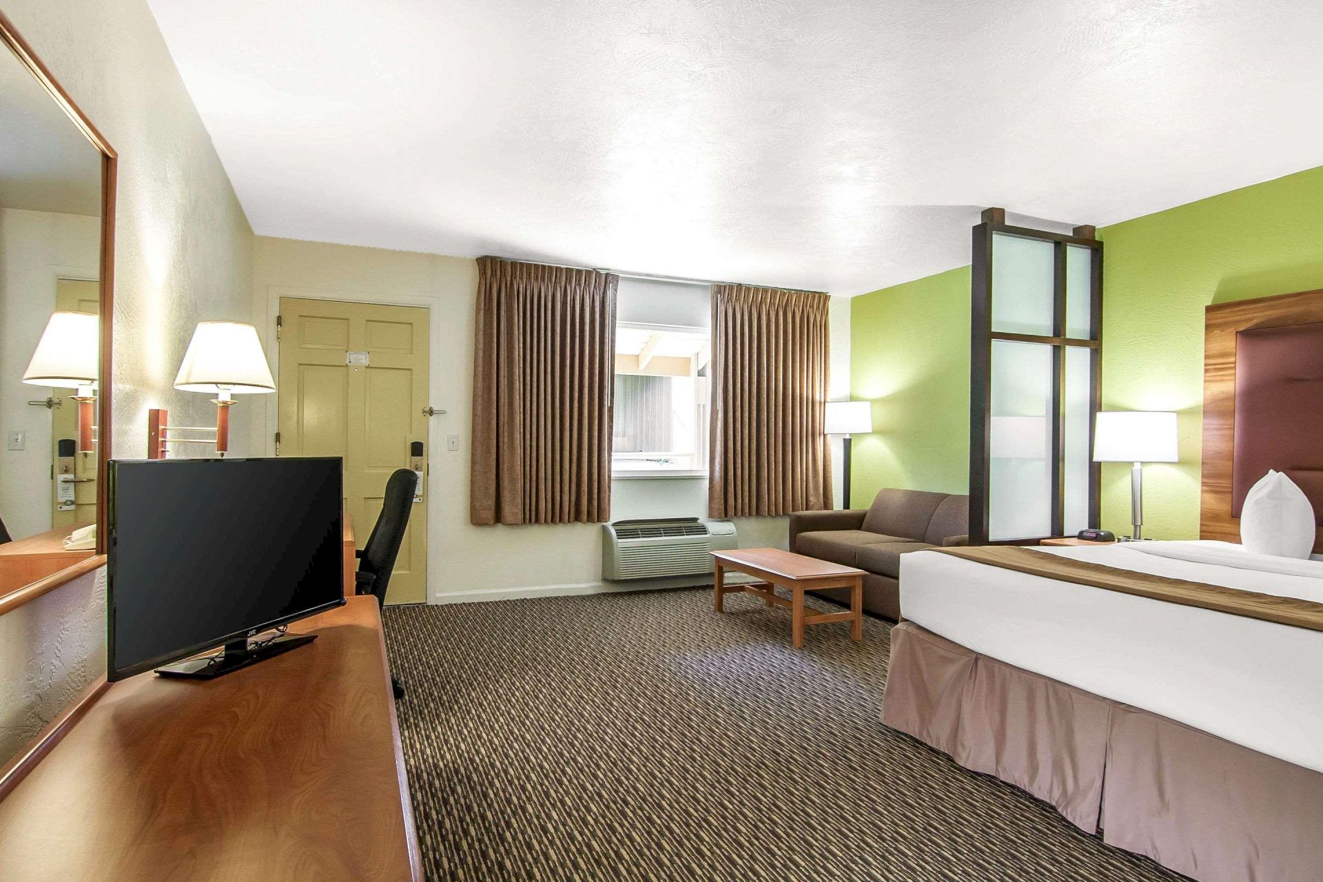 A hotel room with a bed, TV, sofa, and table. It has a green accent wall and warm lighting, creating a cozy and inviting atmosphere.