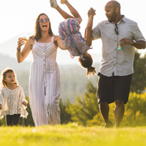 A family of four enjoys a sunny day outside; parents swing a child in the air while another child walks alongside, all smiling and having fun.