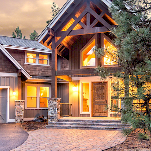 A large house with lit windows, surrounded by trees, featuring a two-car garage and a stone pathway leading to the entrance at dusk.