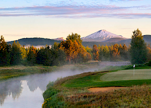 A serene landscape featuring a golf course beside a tranquil river with a snow-capped mountain and lush forests in the background, under a clear sky.