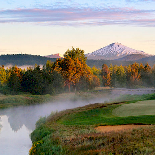 A serene landscape featuring a golf course beside a tranquil river with a snow-capped mountain and lush forests in the background, under a clear sky.