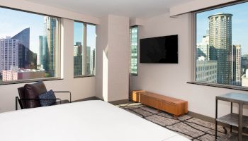 A modern hotel room with large windows showcasing a city view, a wall-mounted TV, a chair, and a bench present in the room.