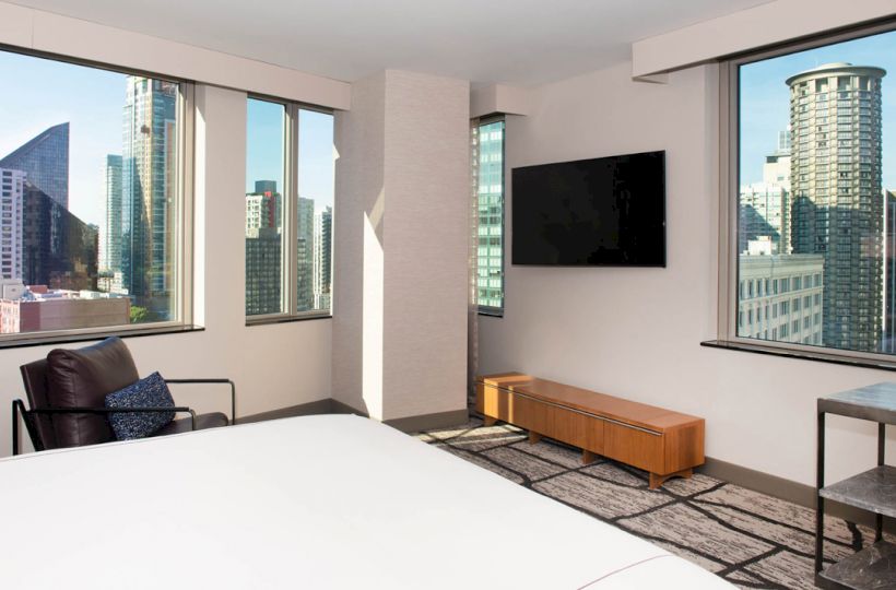 A modern hotel room with large windows showcasing a city view, a wall-mounted TV, a chair, and a bench present in the room.