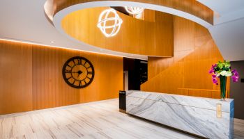 A modern lobby with a marble reception desk, wooden walls, a large clock, and floating light fixtures. There's a vase with flowers on the counter.