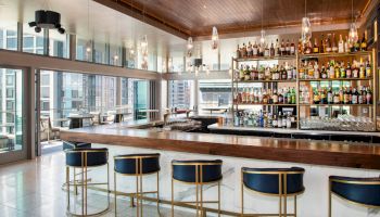 The image shows a modern bar with a wooden counter, blue-gold bar stools, hanging lights, large windows, and a wide selection of liquor bottles on shelves.