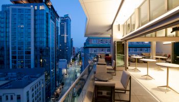 A modern rooftop terrace with chairs and tables overlooking a city skyline at dusk, with tall buildings and illuminated windows visible.