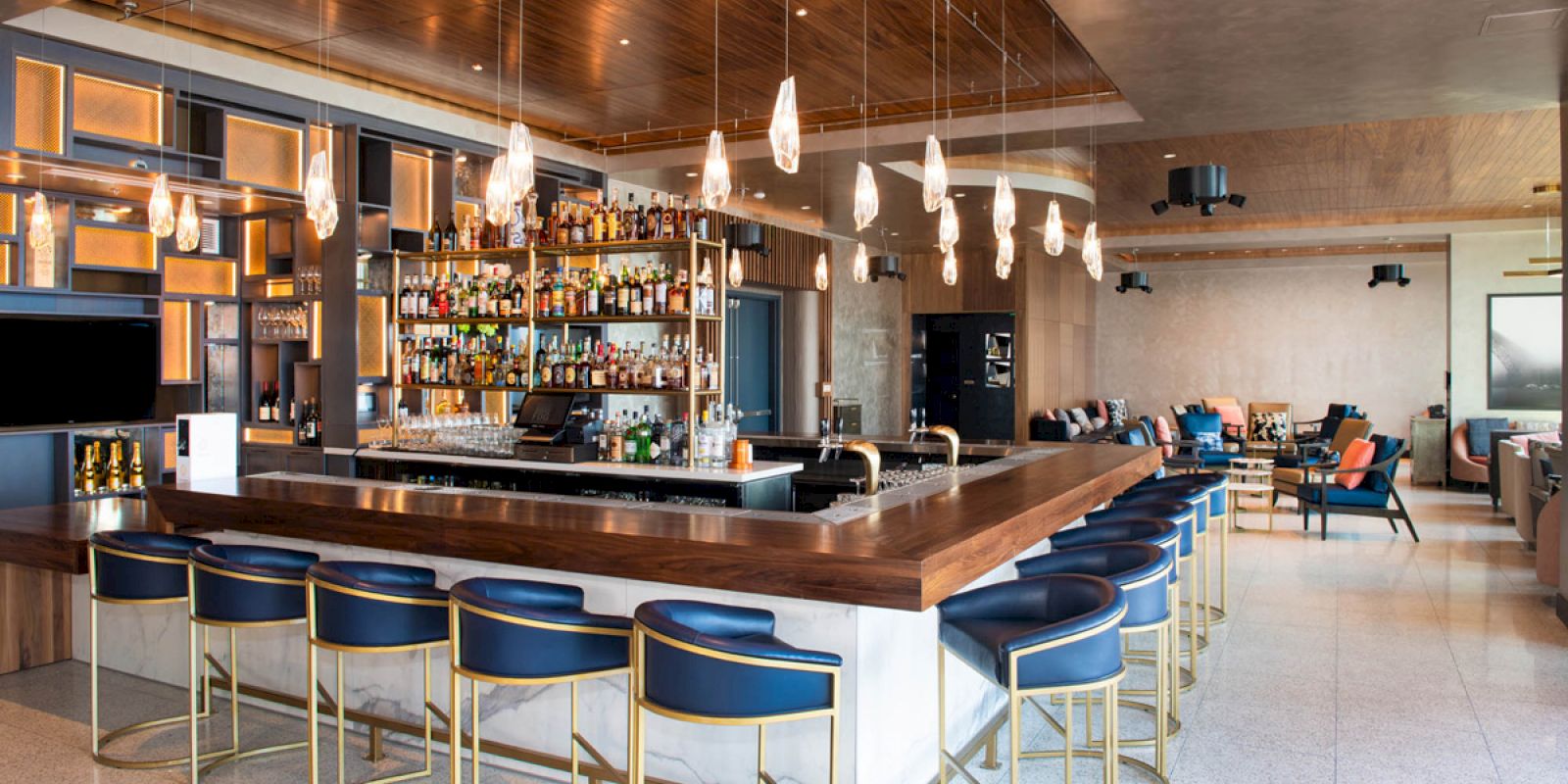 A modern bar with blue stools, wooden counter, glass shelves of liquor, and hanging lights in a spacious lounge setting.