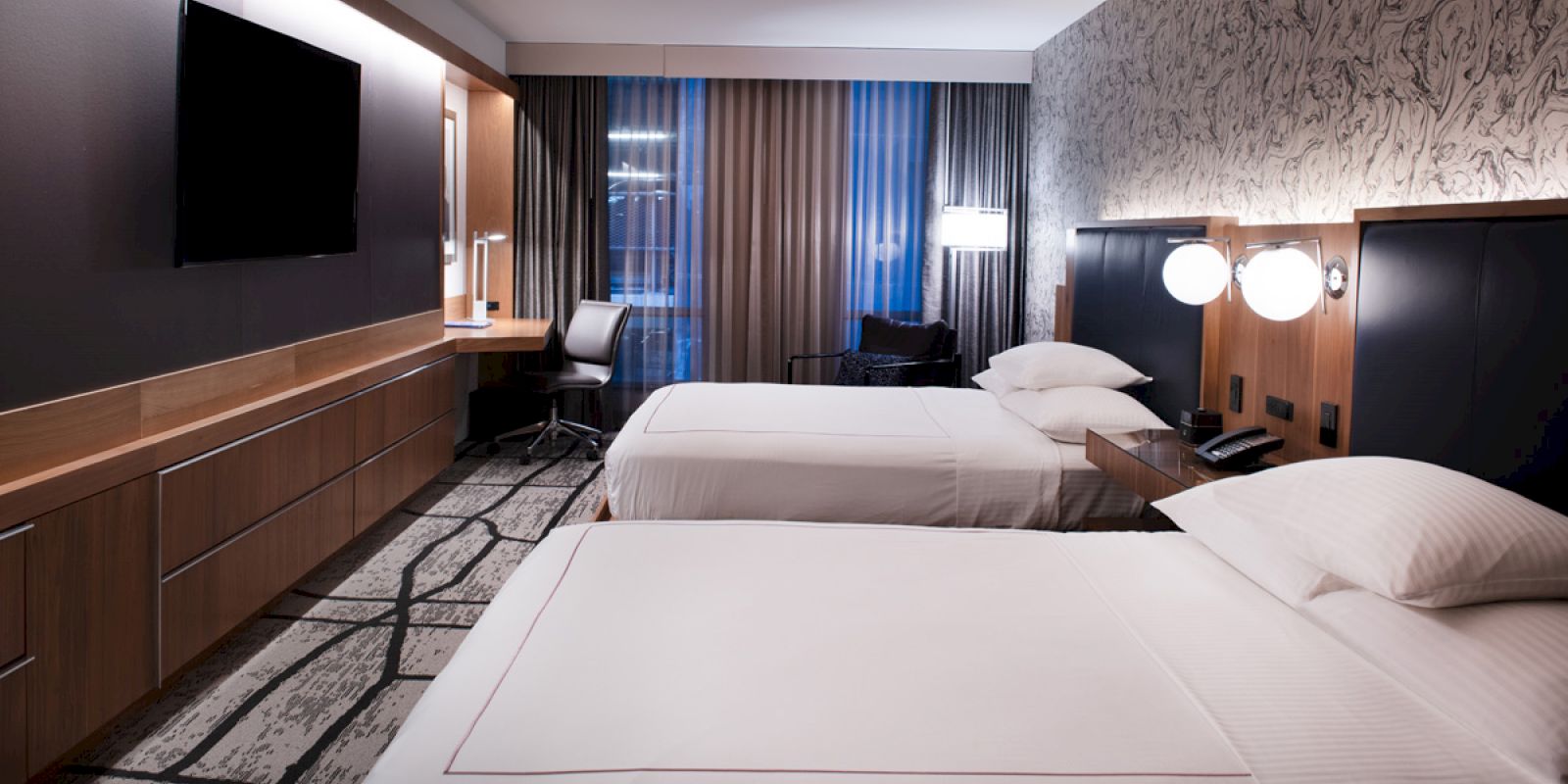 A modern hotel room with two beds, a TV, a work desk, a chair, and stylish decor, illuminated by natural and artificial light.