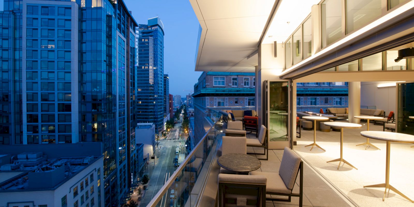 A modern cityscape at dusk with tall buildings, viewed from a balcony with tables and chairs, and a brightly lit interior space.