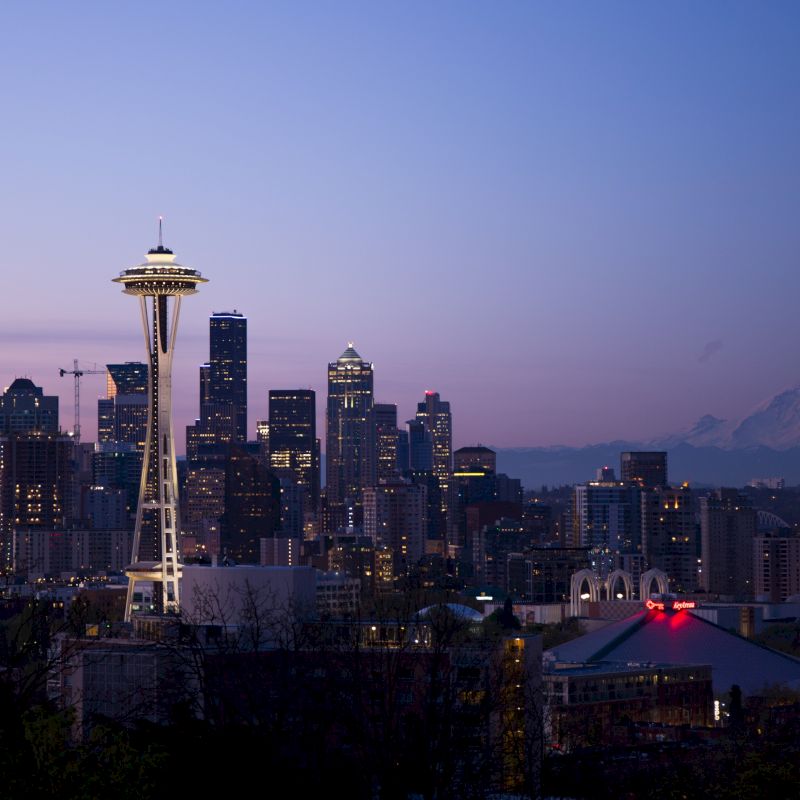 This image shows a city skyline at dusk featuring a prominent tower, with mountains in the background and lights starting to illuminate the city.