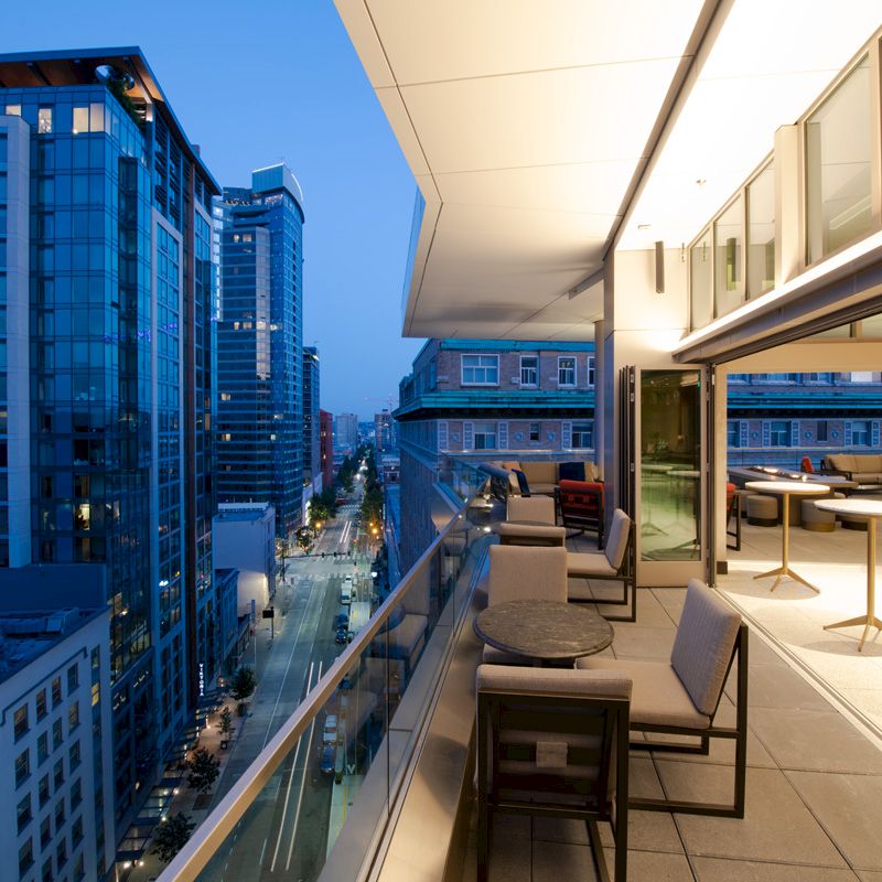 This image shows a modern outdoor balcony at dusk with seating and tables. In the background, there are tall buildings and a city street.