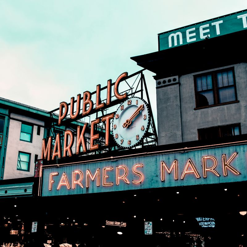 Outdoor market scene featuring "Public Market" and "Farmers Market" neon signs on buildings, with a large clock displayed centrally.