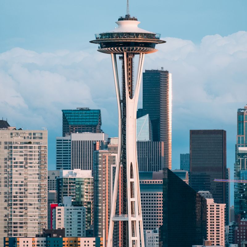 A cityscape with the iconic Space Needle towering over various buildings in Seattle, Washington, under a blue sky with some clouds in the background.