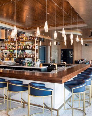 This image shows a modern bar with a wooden counter, blue barstools, hanging light fixtures, and a well-stocked back bar. There’s seating in the background.