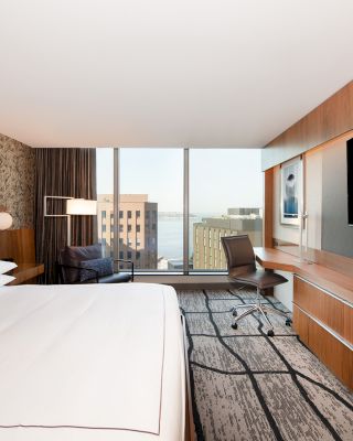 A modern hotel room with a large bed, desk, chair, and flat-screen TV, plus a window with a city view.
