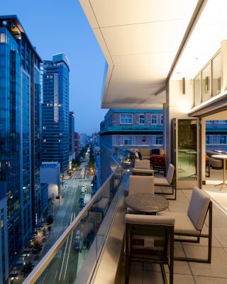 A modern urban balcony overlooks a cityscape with tall buildings and a street below. The lit balcony has chairs and tables, extending indoors.