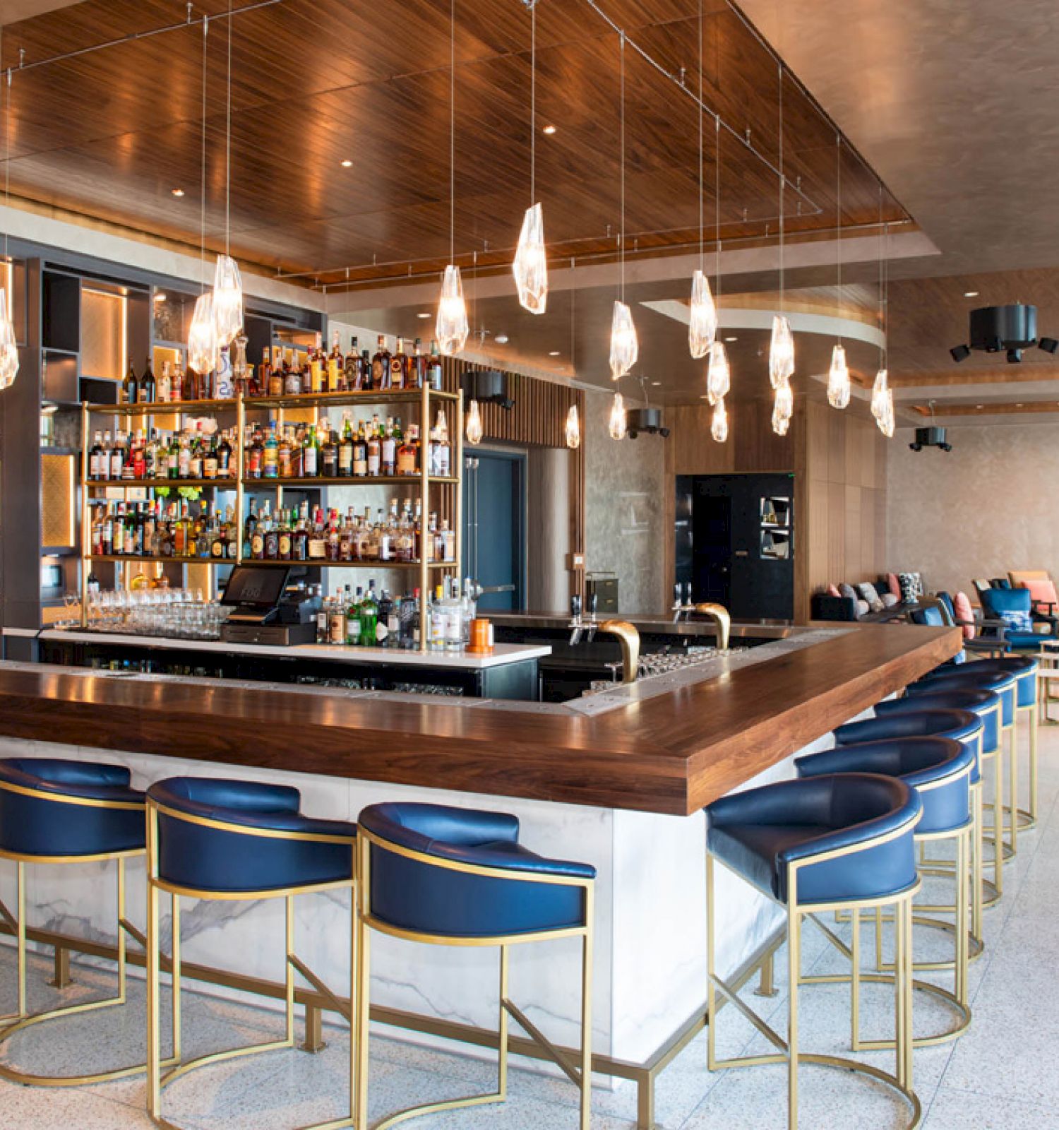 A modern bar with a wooden counter, blue barstools, a well-stocked liquor shelf, and contemporary lighting fixtures in a spacious setting.