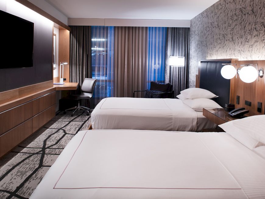 A hotel room featuring two beds, a wall-mounted TV, a desk with a chair by a window, and modern decor with patterned wallpaper and carpeting.