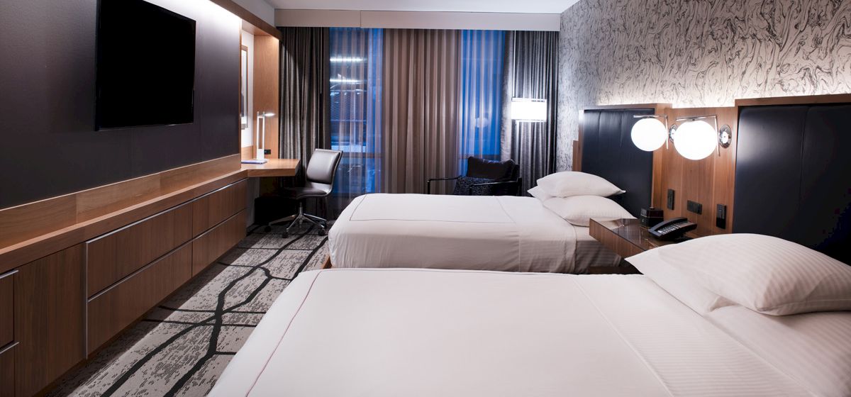 A hotel room featuring two beds, a wall-mounted TV, a desk with a chair by a window, and modern decor with patterned wallpaper and carpeting.
